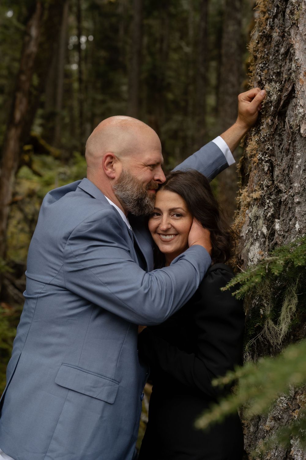 A tender moment shared between a couple against a tree trunk in a forest setting.