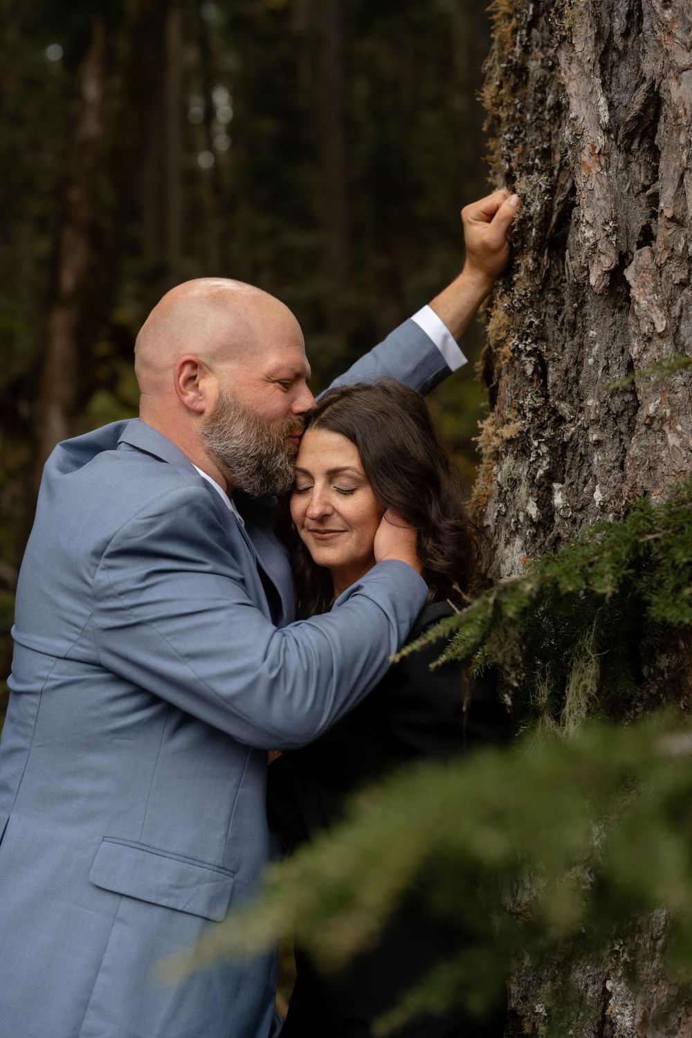 A tender moment between a couple against tall pine trees in a forest setting.