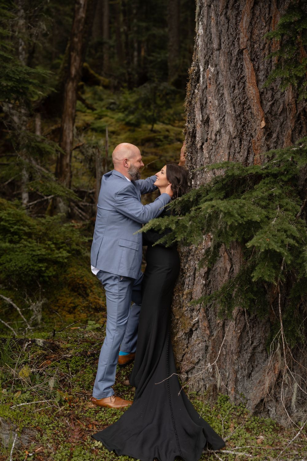 A couple embraces against a large tree trunk in a moody forest setting.