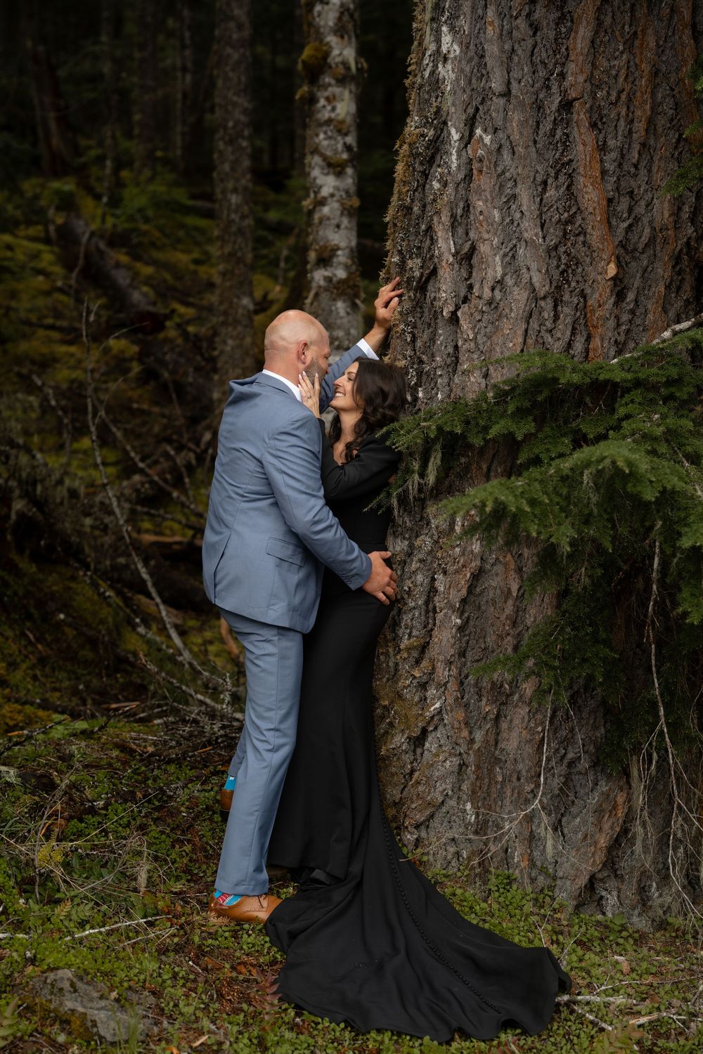 A couple shares an intimate moment against a large tree trunk in a forest setting.