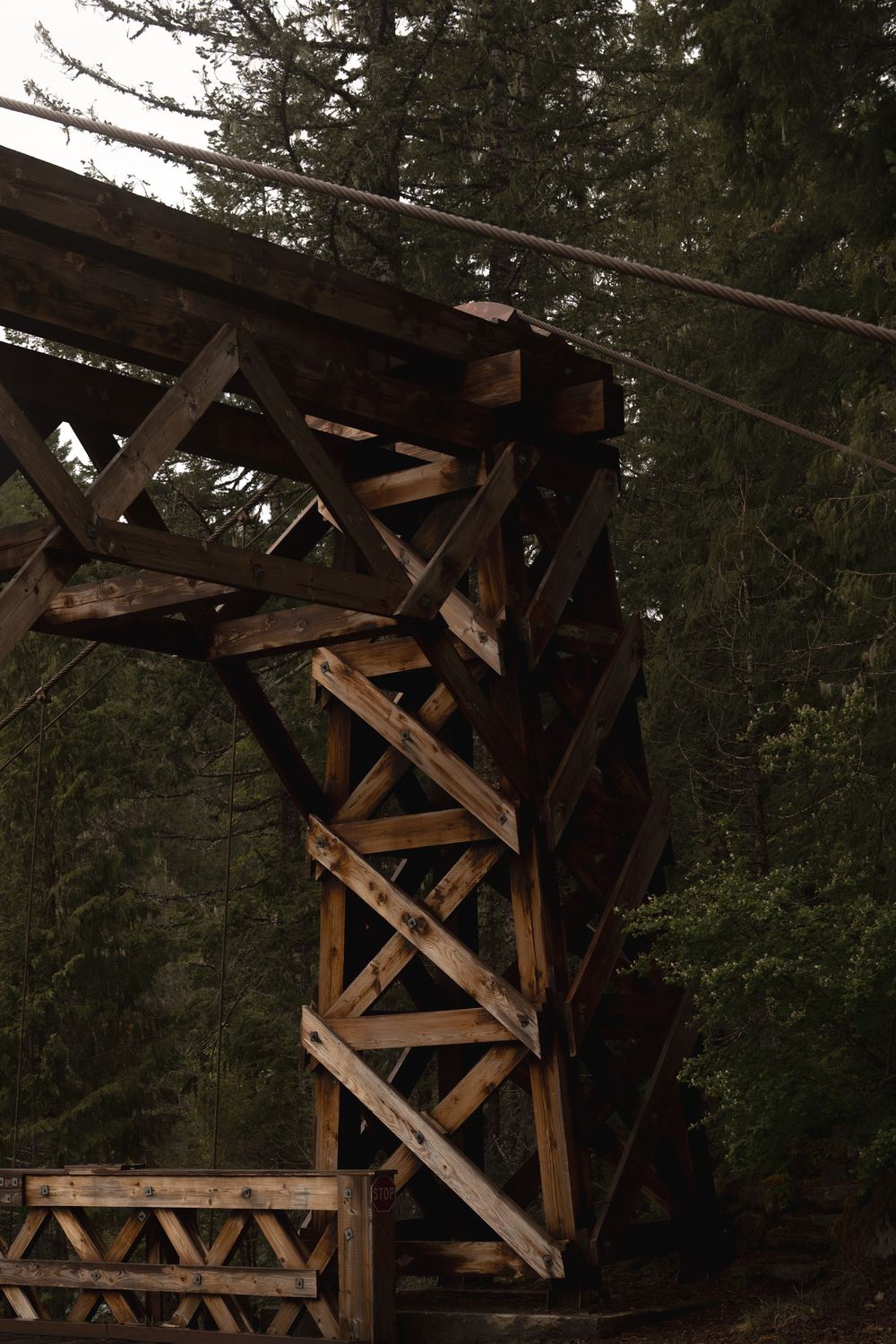Wooden trestle bridge structure with diagonal support beams against a dark forest background.