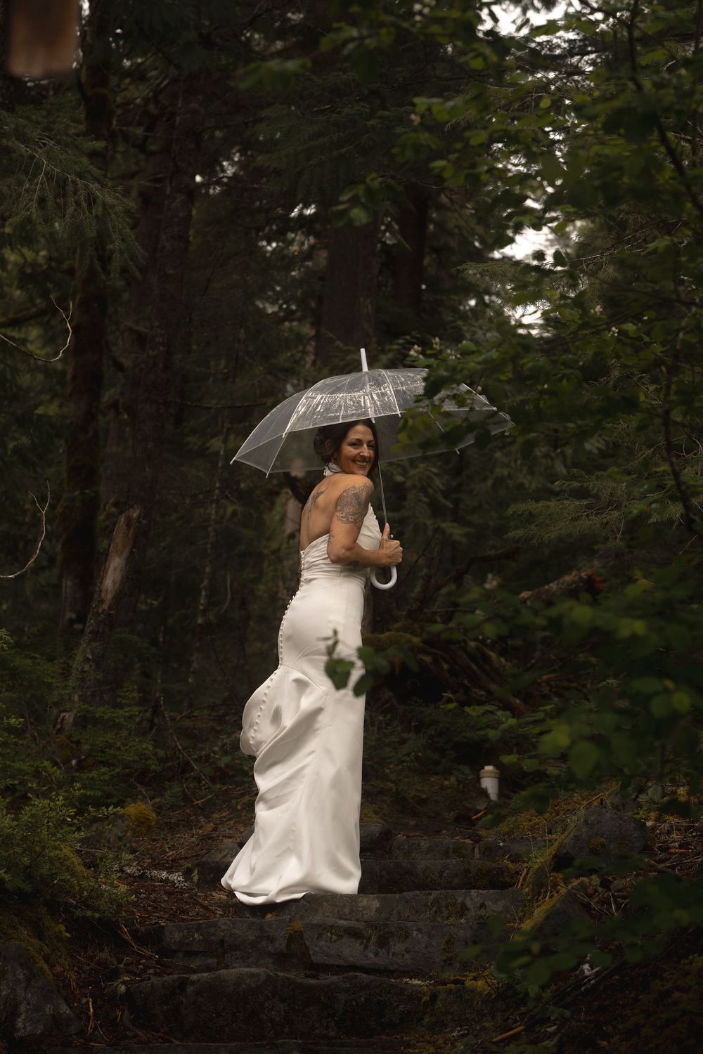 A bride in a white fitted gown poses with a clear umbrella in a misty forest setting.