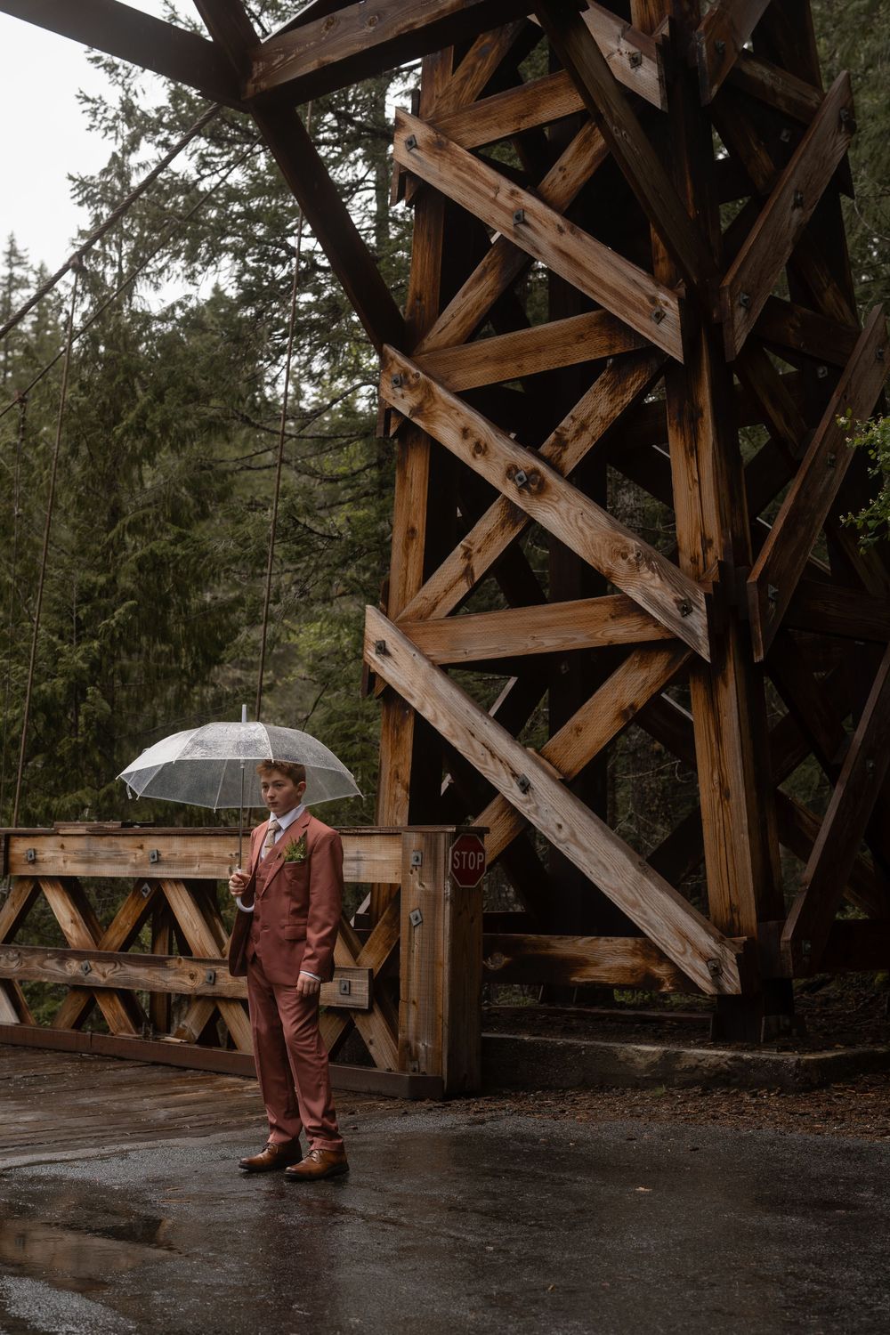 A person with an umbrella stands under a rustic wooden structure in the rain.