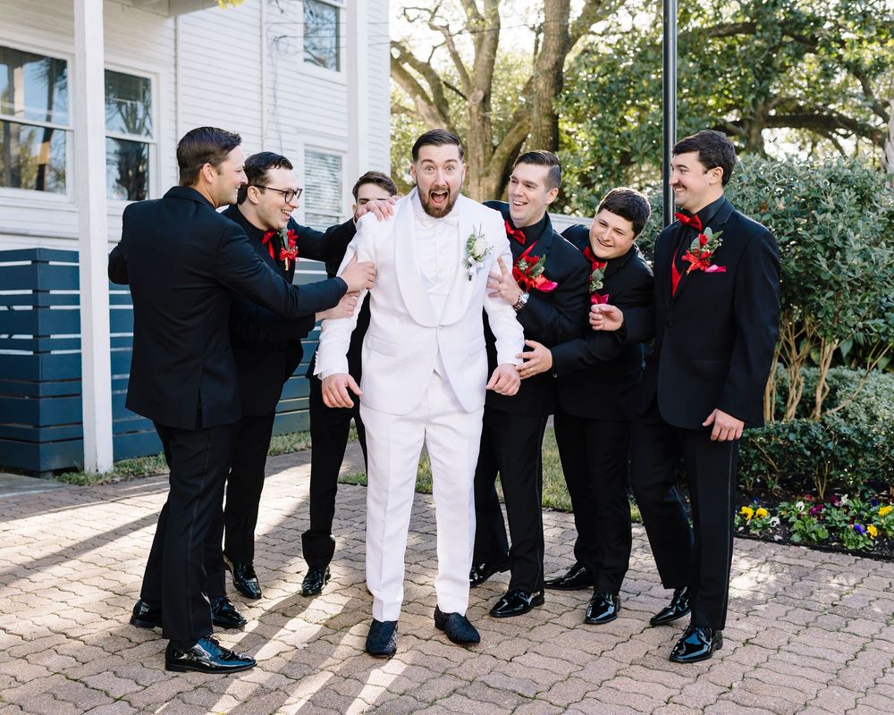 A groom in white suit stands with groomsmen in black suits sharing laughs outside a building.