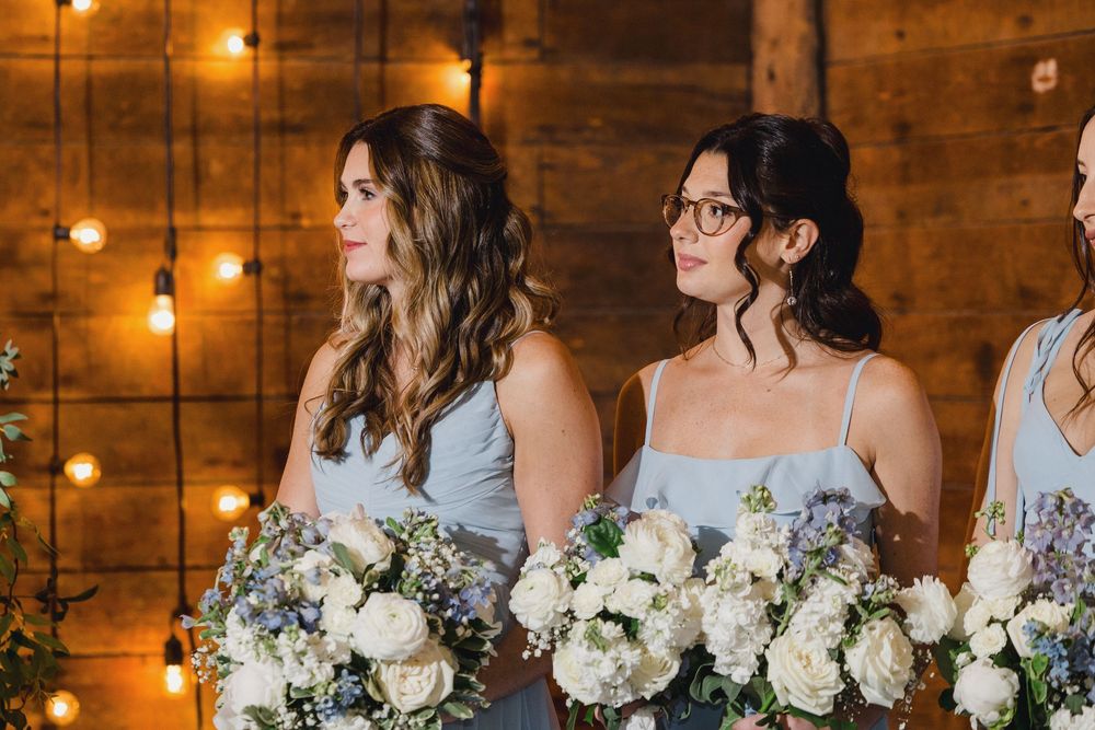 Wedding party members hold white and blue floral bouquets while standing in a rustic venue with warm lighting.