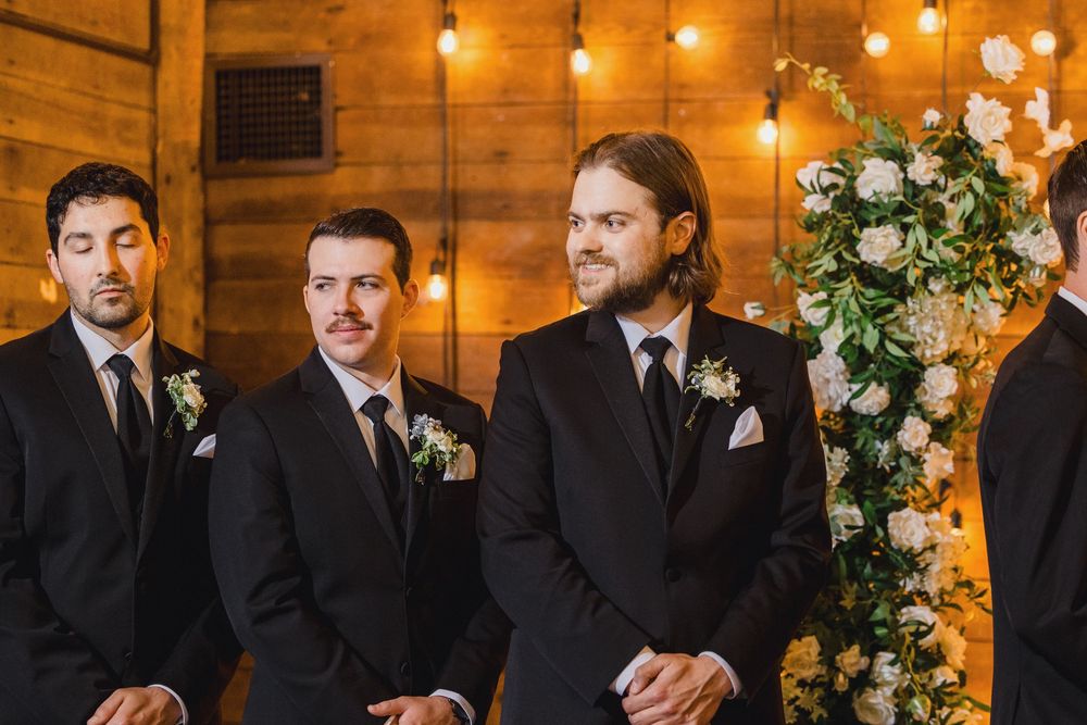 Groomsmen in black suits stand at rustic wooden wedding altar decorated with string lights and floral arrangements.