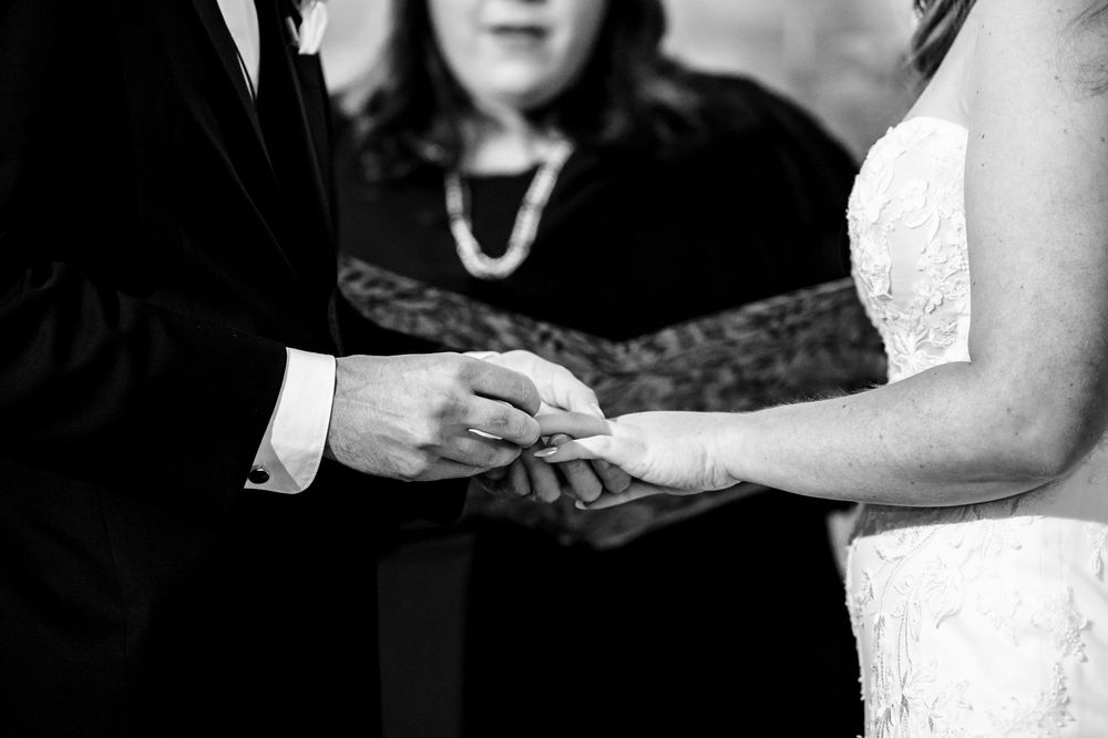 Black and white close-up of hands exchanging wedding rings during a ceremony with officiant in background.