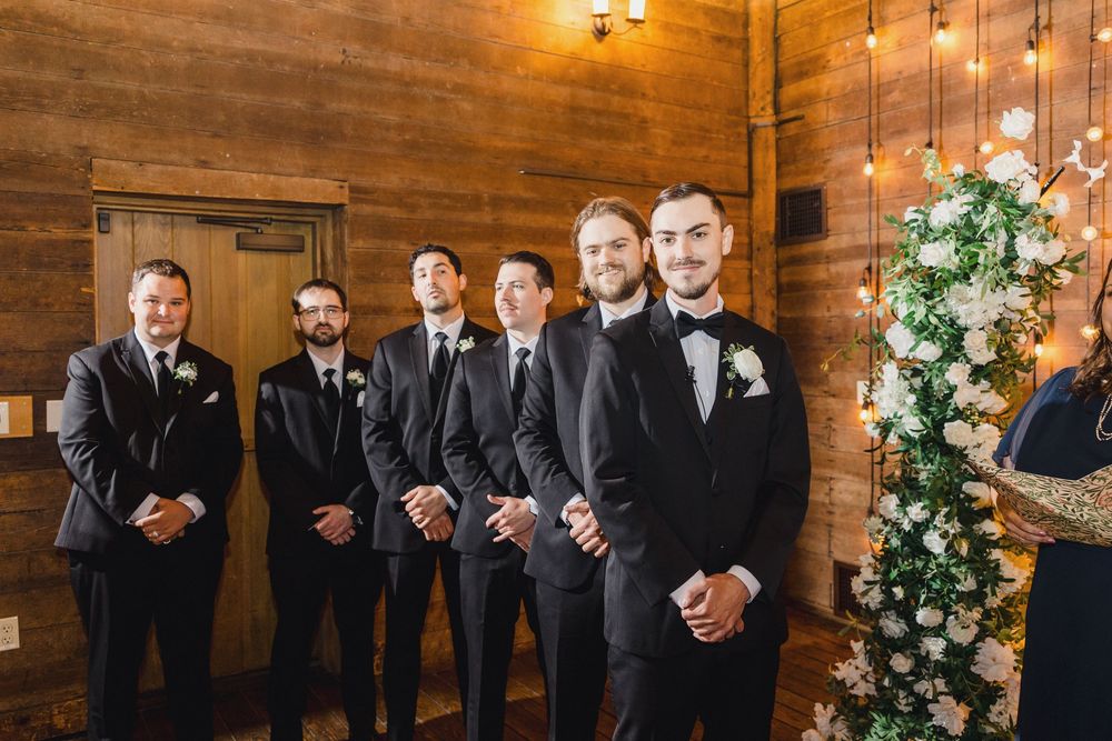 Wedding party members in dark suits pose together inside rustic wooden venue with warm lighting and floral decor.