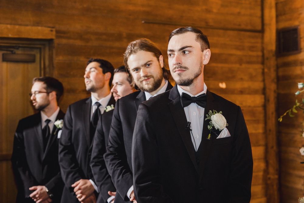 Groomsmen in black suits pose together against rustic wooden wall at wedding venue.