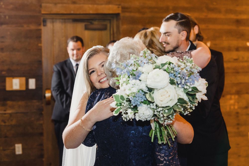 Joyful wedding party poses with beautiful blue and white floral bouquets in rustic wooden venue.