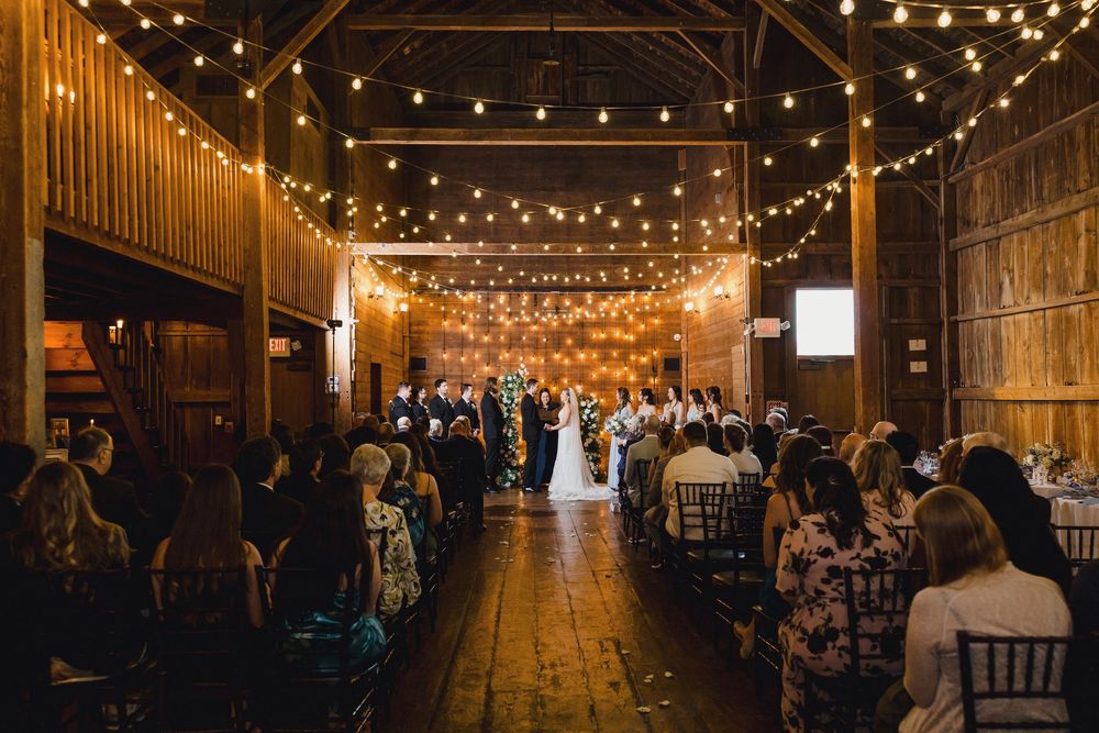 String lights illuminate a rustic barn wedding reception with guests seated at long wooden tables.