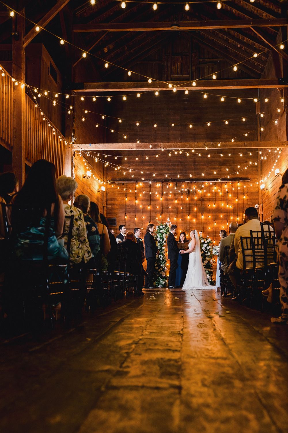 String lights illuminate a rustic barn wedding ceremony with wooden beams and warm ambient lighting creating a romantic atmosphere.