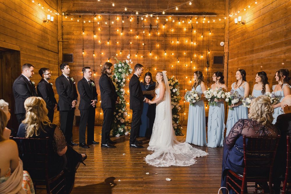 Wedding ceremony in a rustic wooden barn with string lights and floral arch creating a warm, romantic atmosphere.