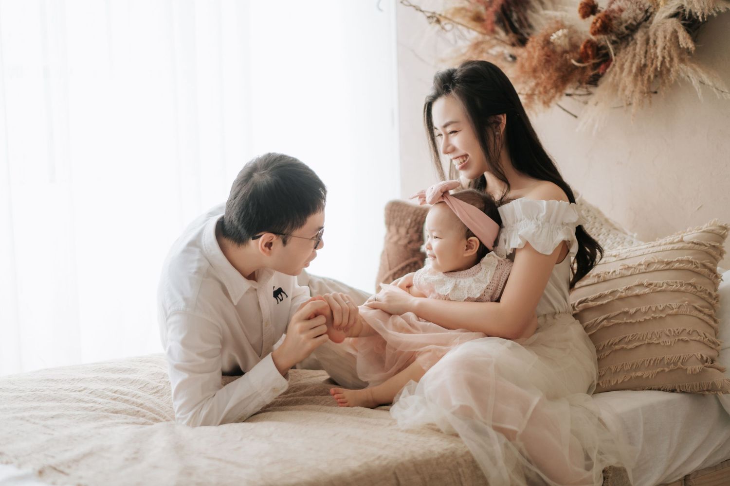 A young family shares a tender moment on a bed, with parents smiling at their baby in a bright, cozy room.一個年輕的家庭在床上享受著溫馨的時刻，父母在明亮舒適的房間裡微笑著看