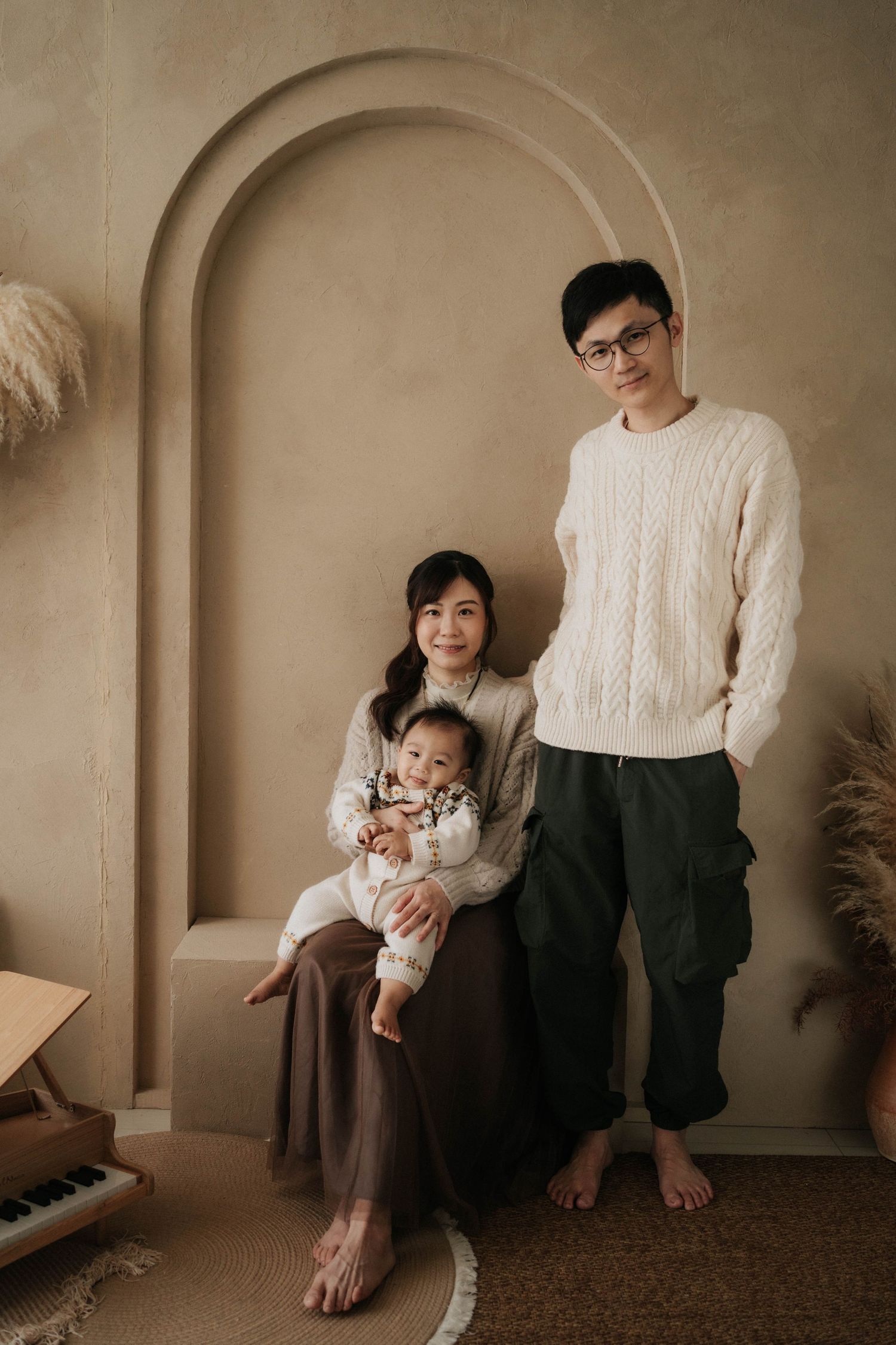A young Asian family poses in a warm beige studio, parents in cream sweaters with their baby, beside a toy piano.一個年輕的亞洲家庭在溫暖的米色攝影棚裡擺姿勢拍照
