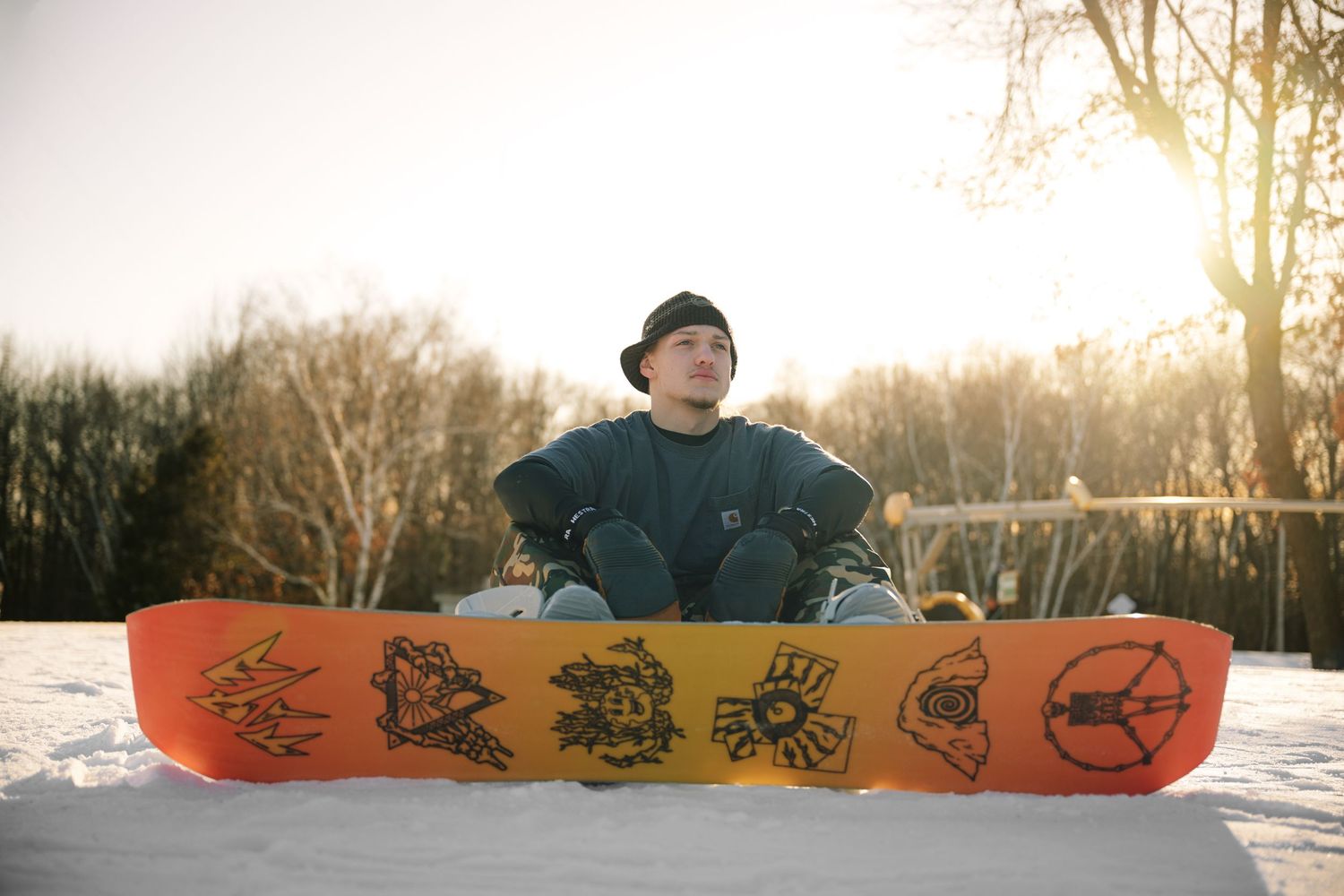 Snowboarder sitting behind colorful orange and red snowboard on snowy slope in bright sunlight.