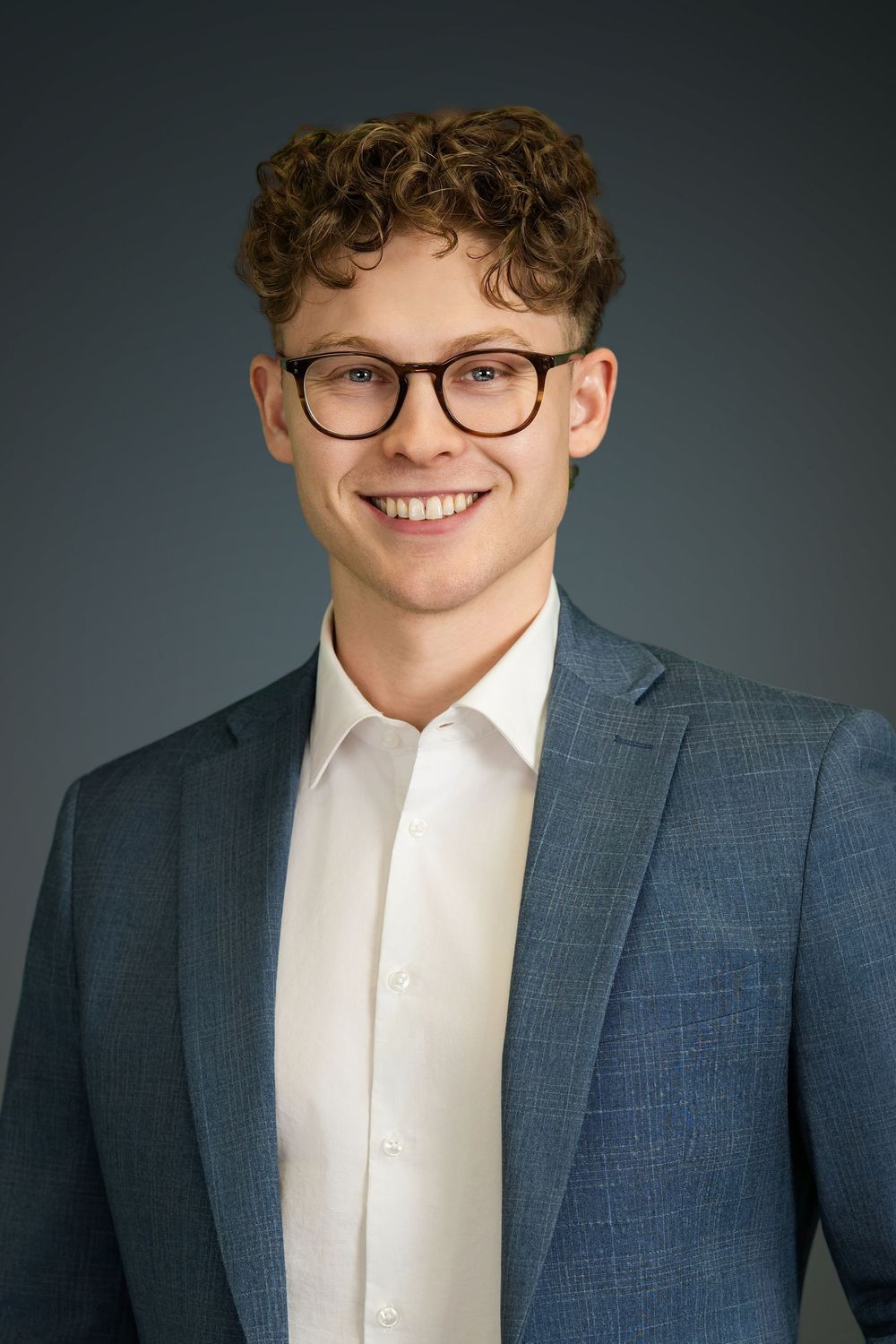 Professional headshot portrait of someone wearing a navy suit jacket, white dress shirt and round glasses against dark backdrop.