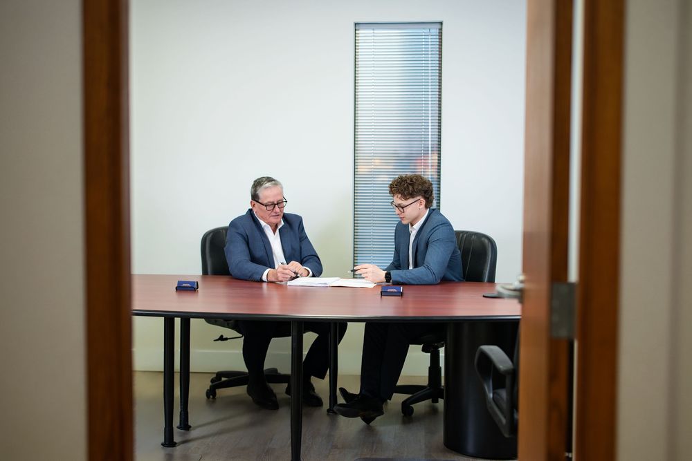 Two professionals in business attire sitting at a desk in a meeting room with glass walls.