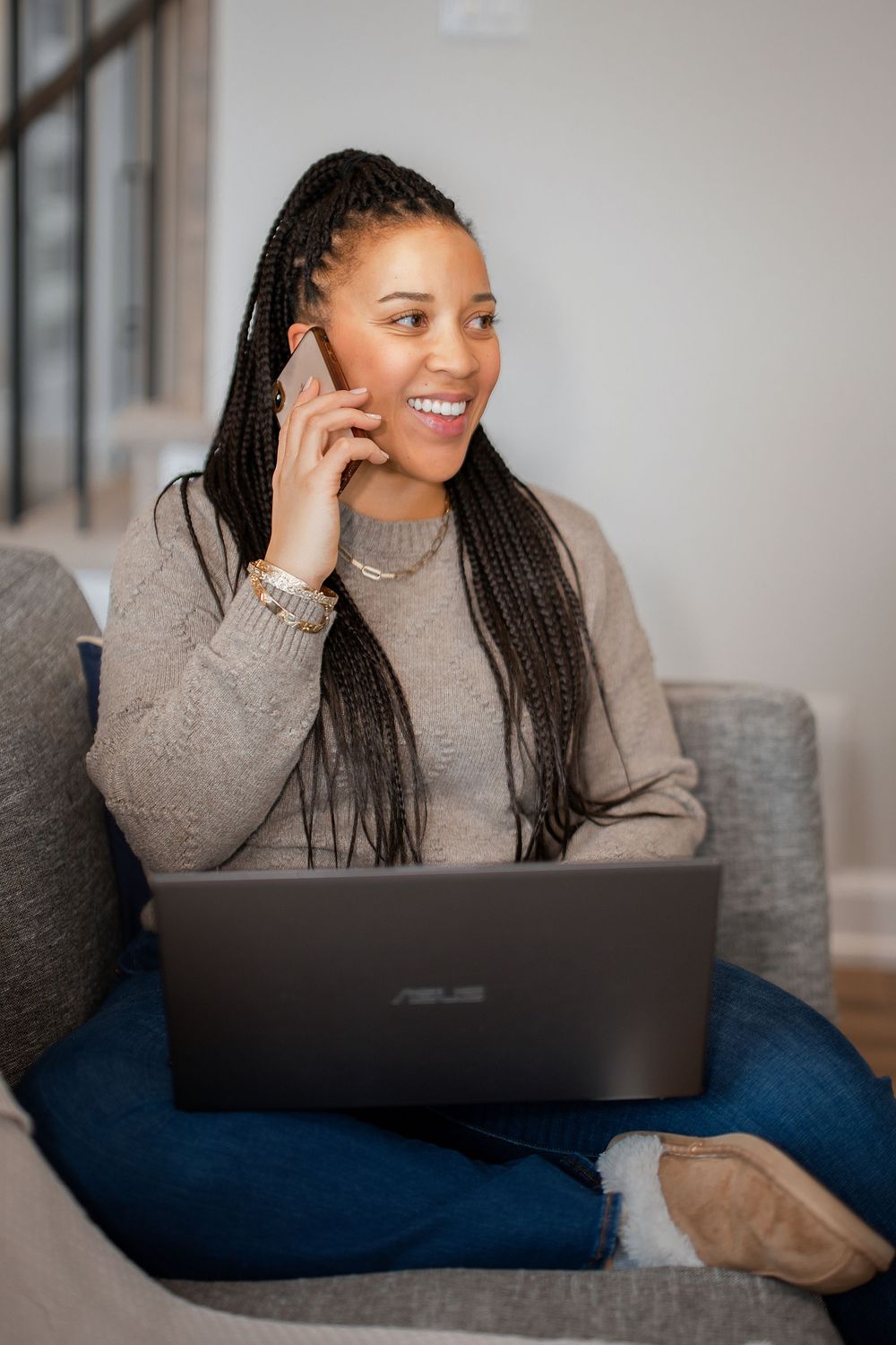 Woman in grey sweater smiles while working on laptop from comfortable couch.