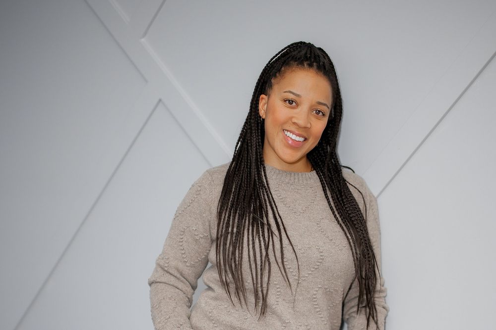 A person with long dark braids poses in a cozy beige turtleneck sweater against a white geometric wall background.