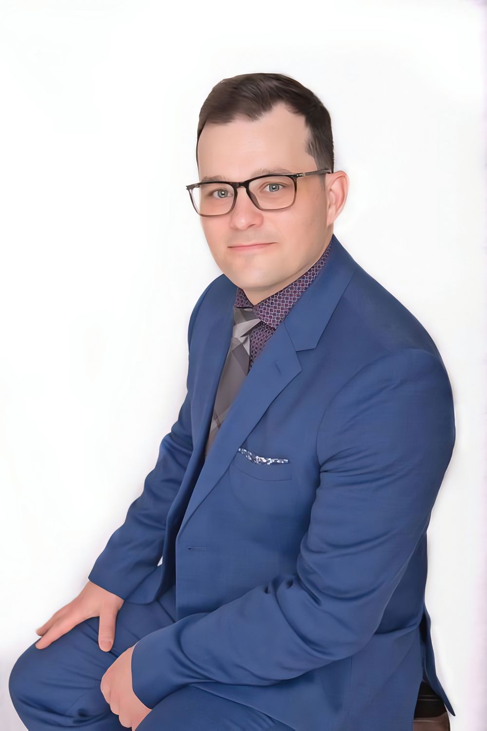 Professional headshot of person in blue suit jacket, patterned shirt and tie against white background.