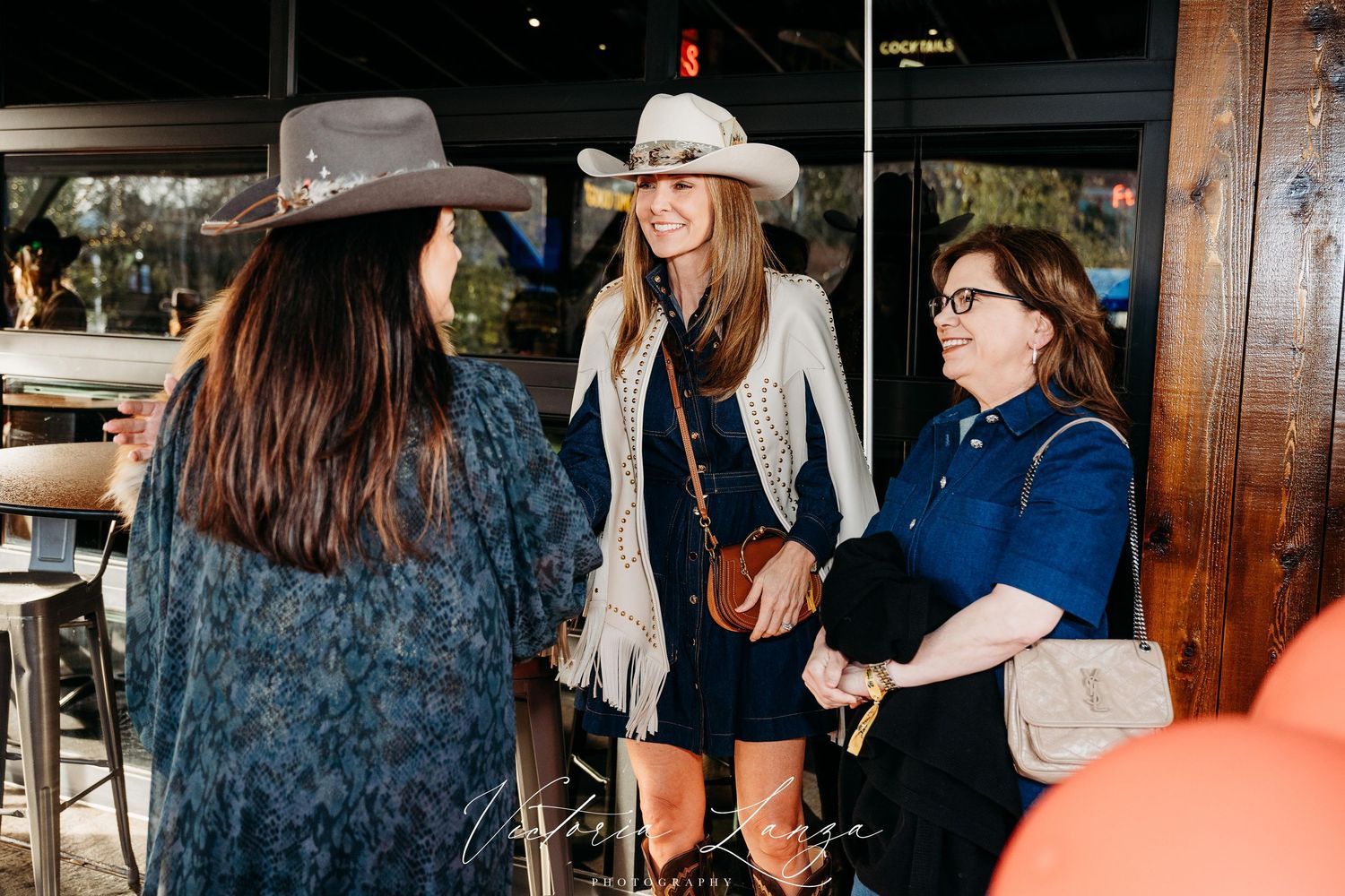 Three people chat together at a casual gathering, wearing western-style hats and casual attire.