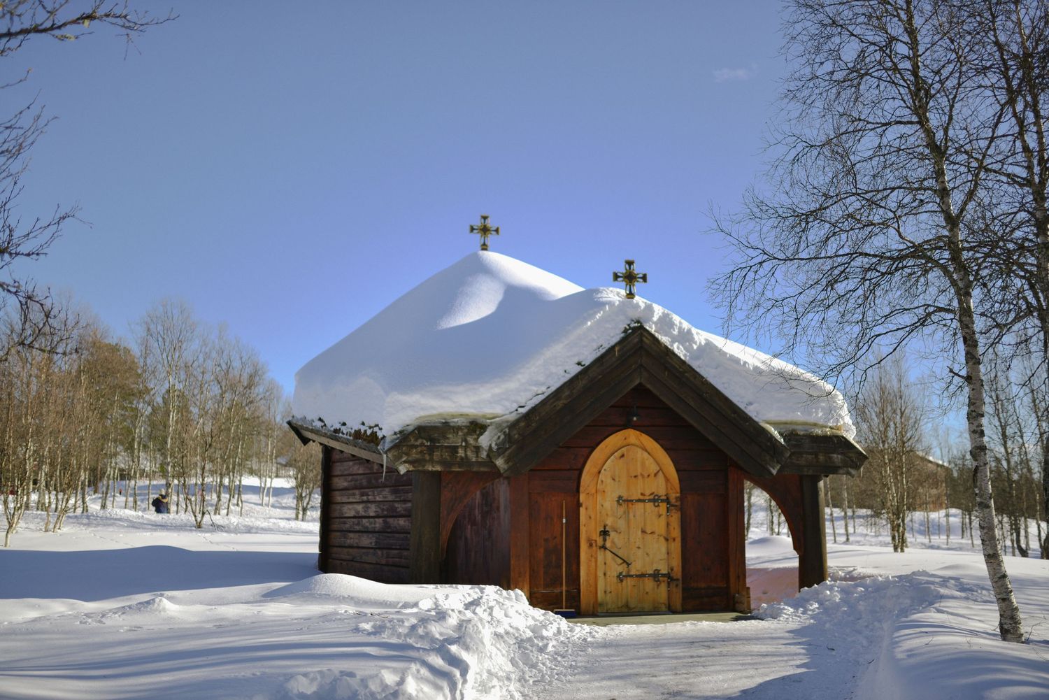 A small wooden chapel with snow-covered roof stands in a winter forest landscape beneath a bright blue sky.