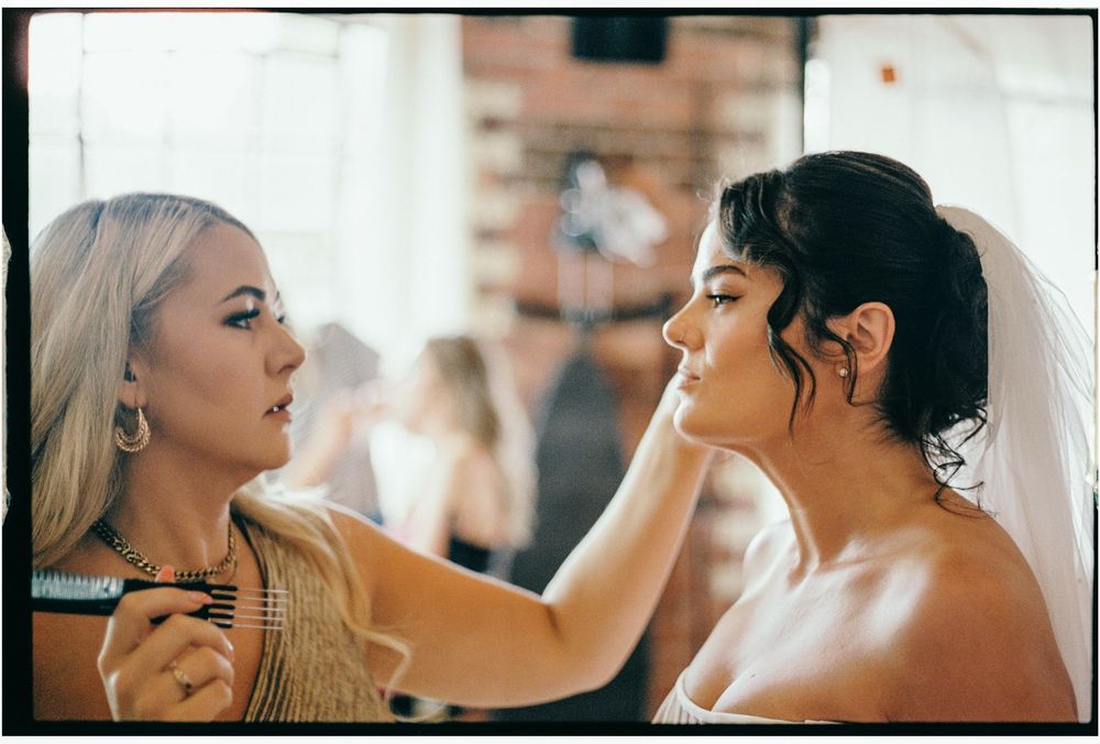 Professional makeup artist applies cosmetics to model's face in studio setting with natural window lighting.