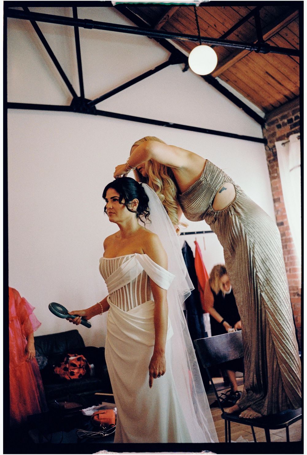 A bride in a white corset dress getting ready with help from a stylist in a beige gown during wedding preparations.
