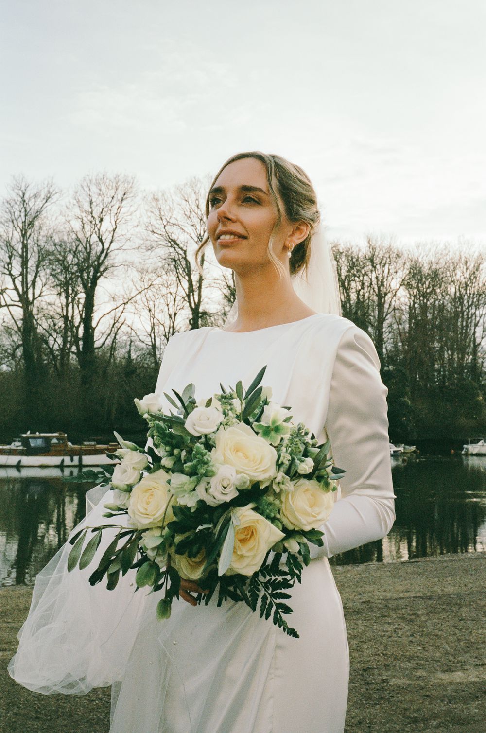 A bride in a long-sleeved winter wedding dress holds a lush white rose bouquet with greenery against a scenic outdoor backdrop.