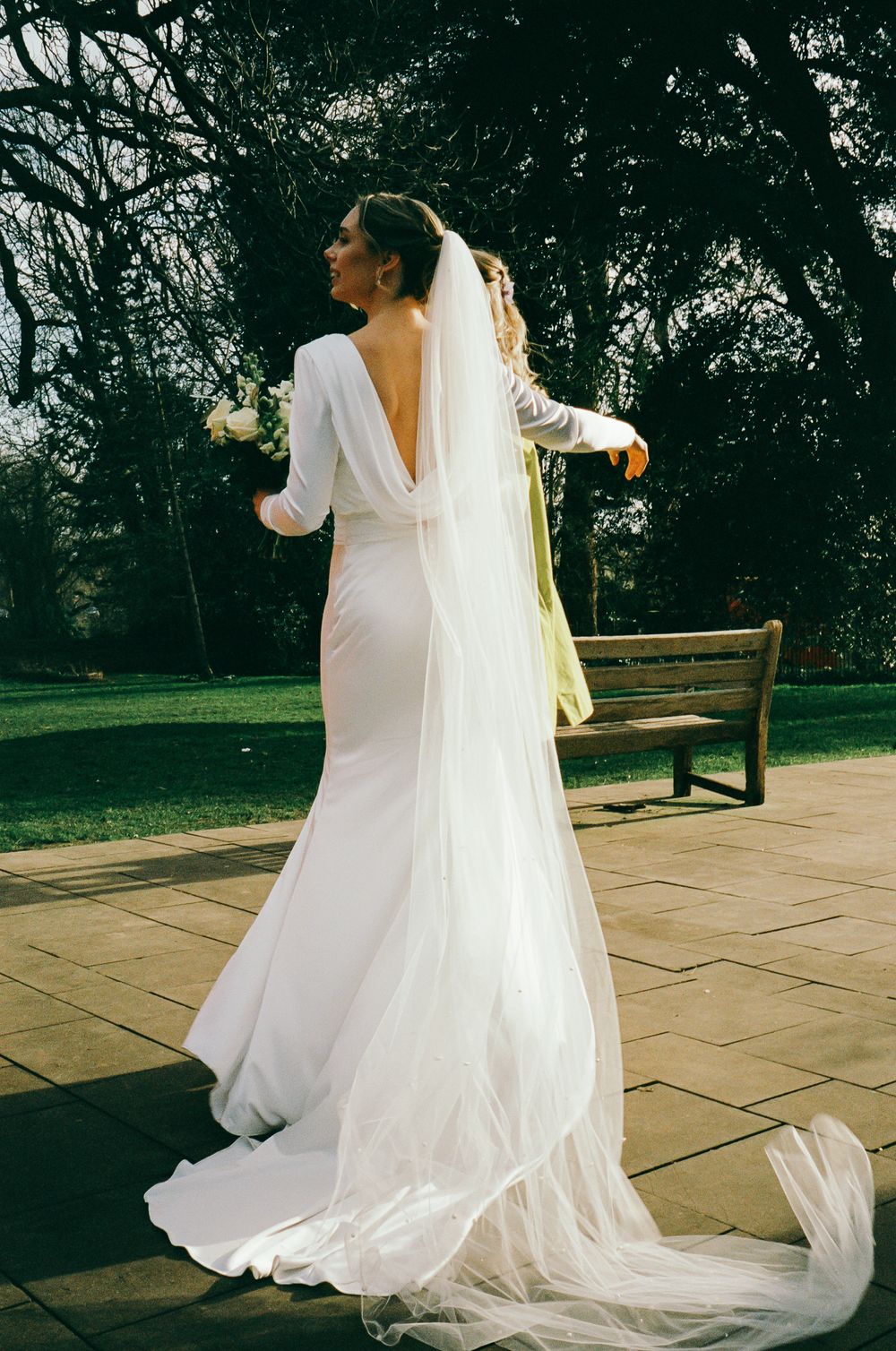 A flowing white wedding dress and veil twirl gracefully on a stone patio surrounded by trees in natural light.