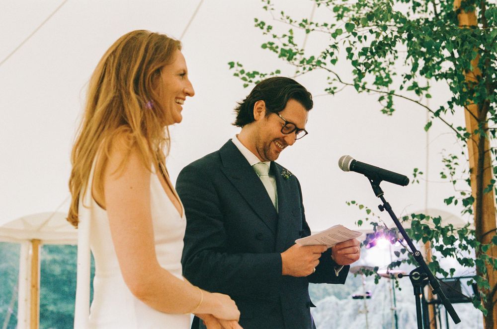 A couple stands at a microphone under a white tent during a wedding celebration.