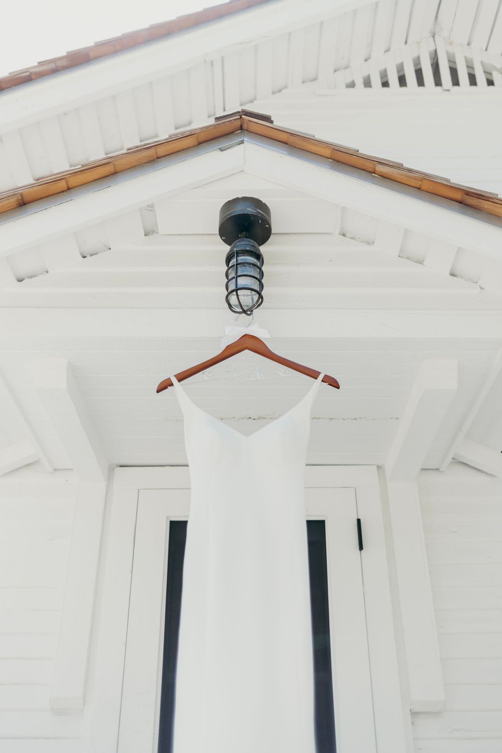 A white sleeveless dress hangs on a wooden hanger against a bright white wall with exposed wooden beams.