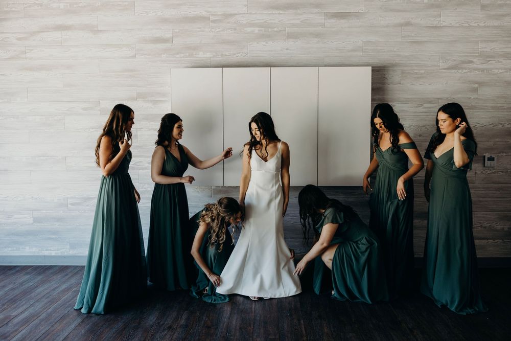 Bridesmaids in teal dresses help adjust a white wedding gown against a gray textured wall.