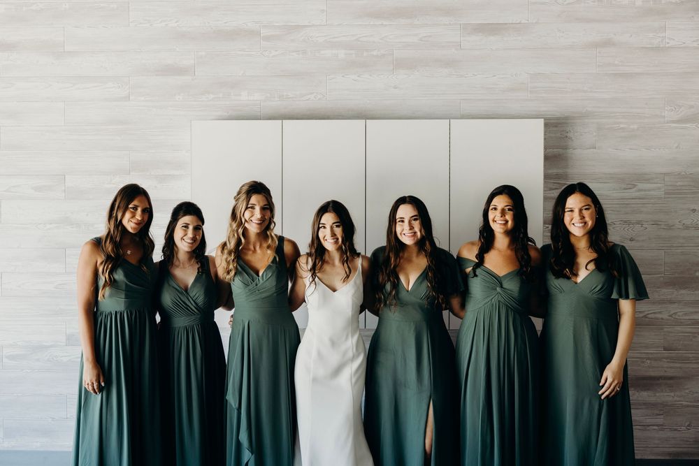 A wedding party wearing sage green bridesmaid dresses poses together against a light gray wall.