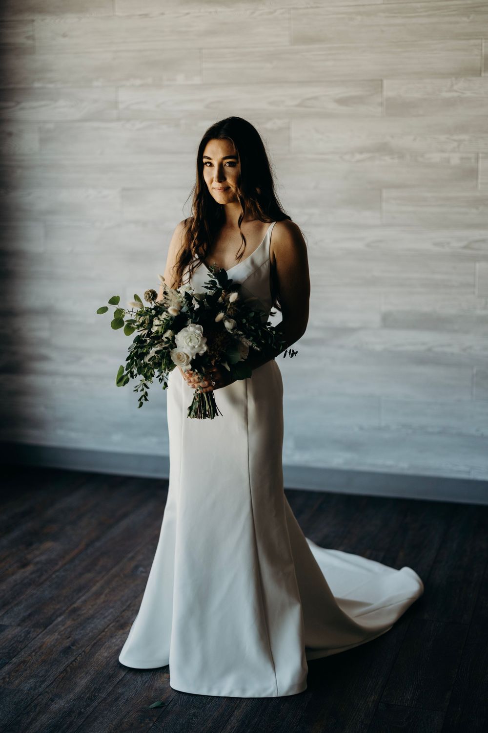 A person in a flowing white dress poses with a lush bouquet against a gray wall in a minimalist setting.