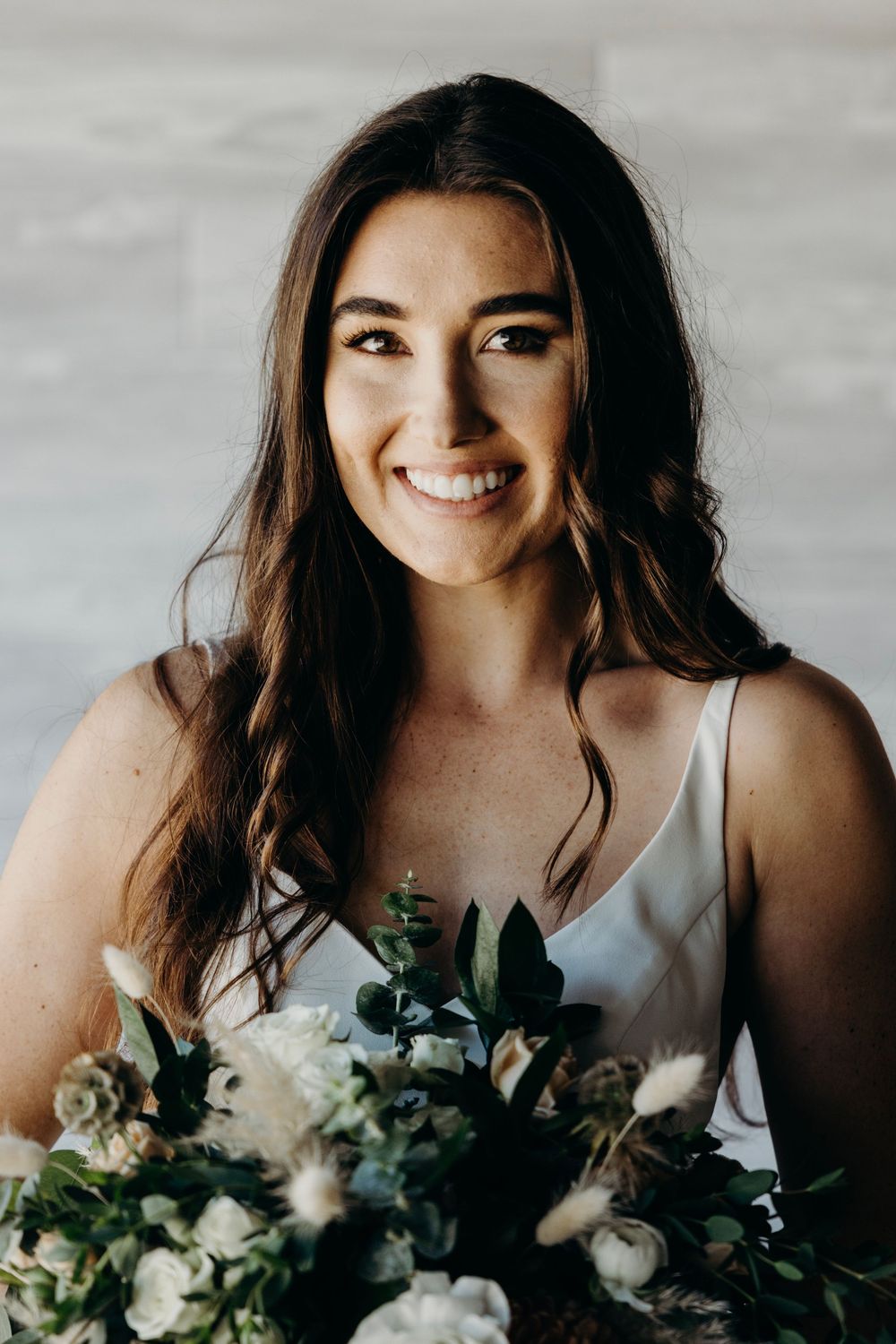 A joyful bride in a white dress holds a lush bouquet of white flowers and greenery against a light background.