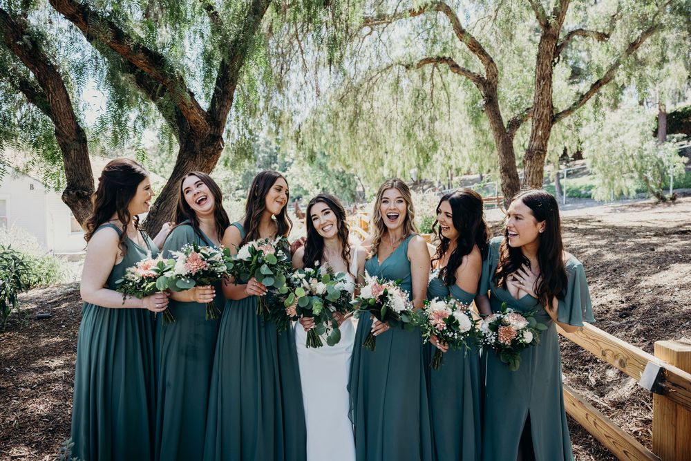 Bridesmaids in elegant emerald green dresses pose together under willow trees at an outdoor wedding celebration.