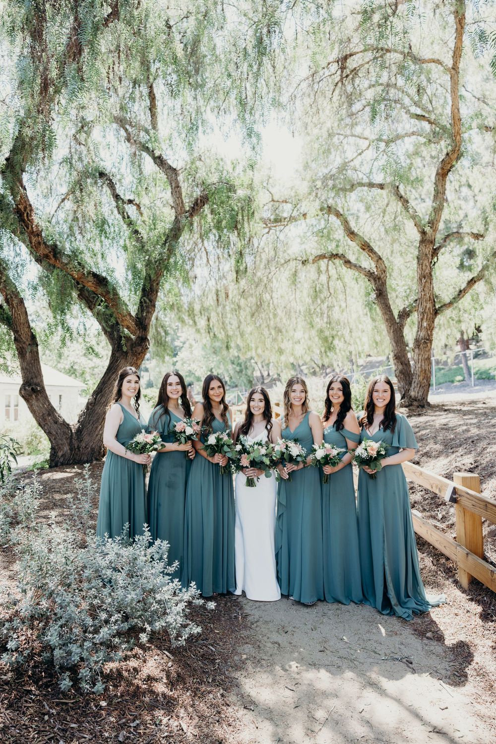 Wedding party in teal bridesmaid dresses poses under drooping willow trees in an outdoor garden setting.