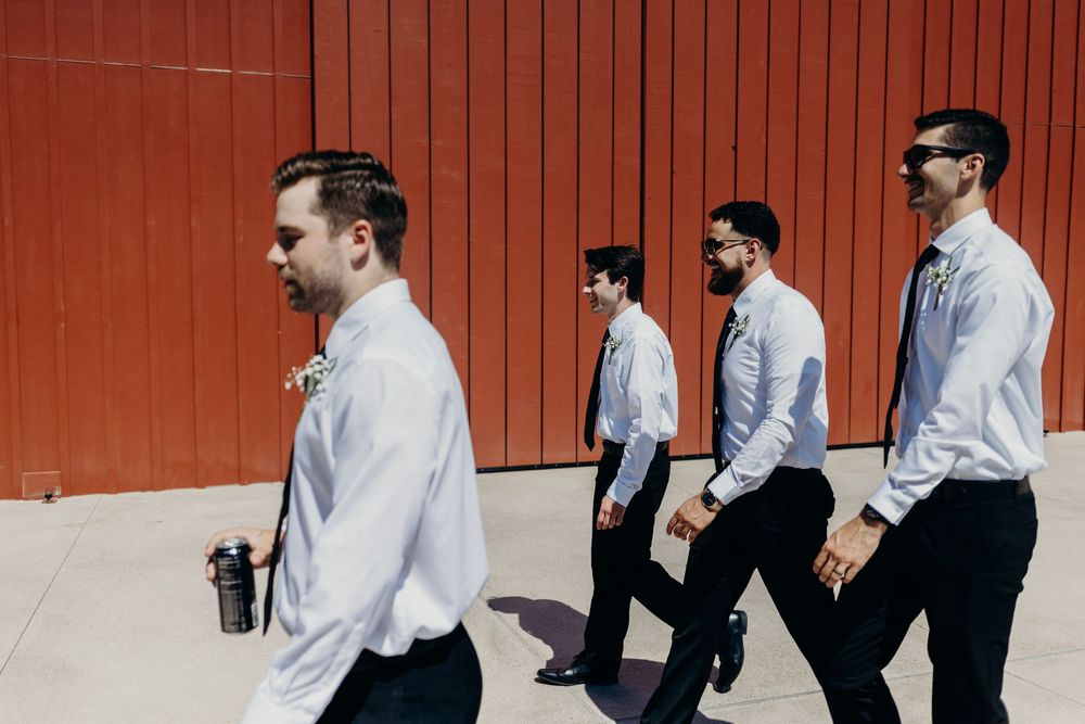 Four men in white dress shirts and black pants walking in a line against an orange wall.