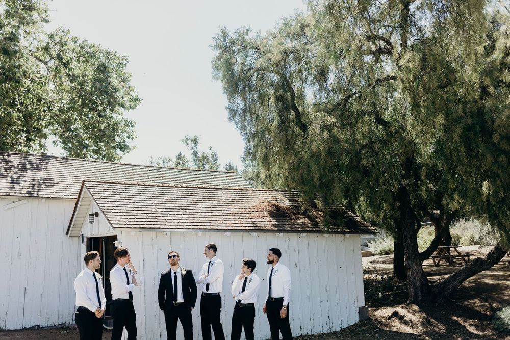 A group in formal attire stands together outside a rustic white barn building with willow trees in the background.