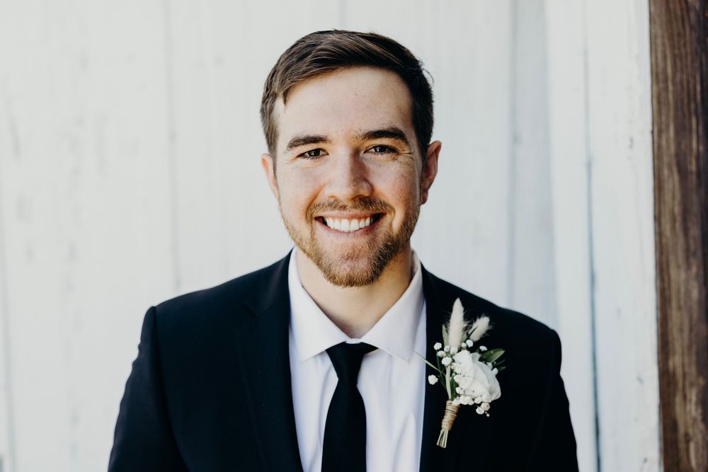 A groom in a black suit and tie smiles warmly while standing in front of a light-colored backdrop.