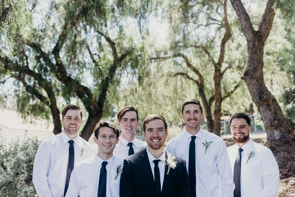 Groomsmen in white shirts and dark ties gather for a photo under willow trees at an outdoor wedding venue.