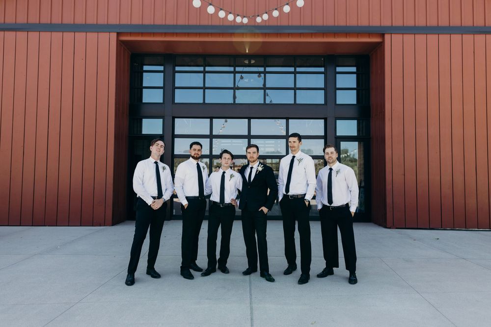 Groomsmen in black suits stand against a red wall for a wedding photo shoot.