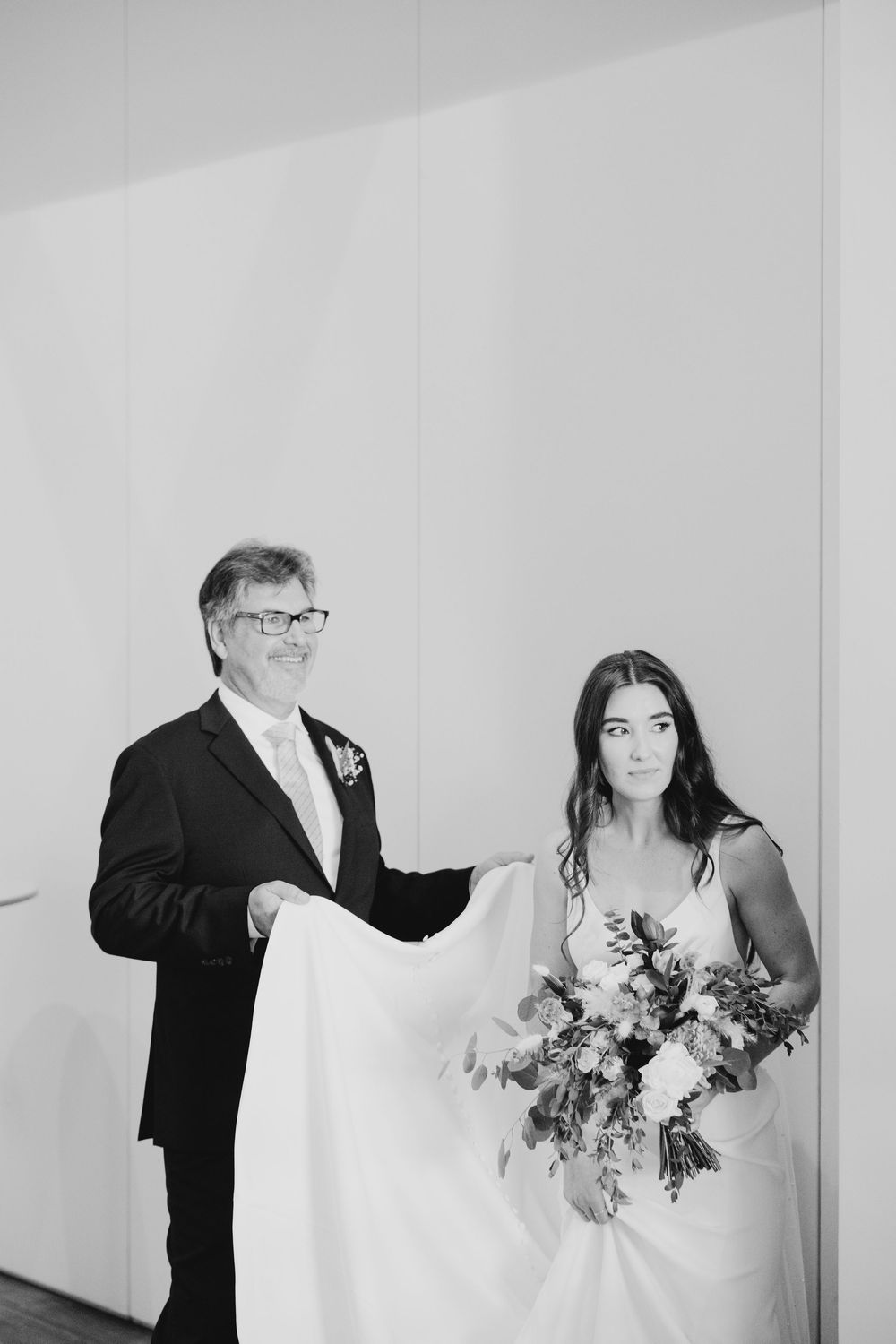 Black and white wedding photo showing a bride in white dress holding flowers with companion in formal suit and glasses.