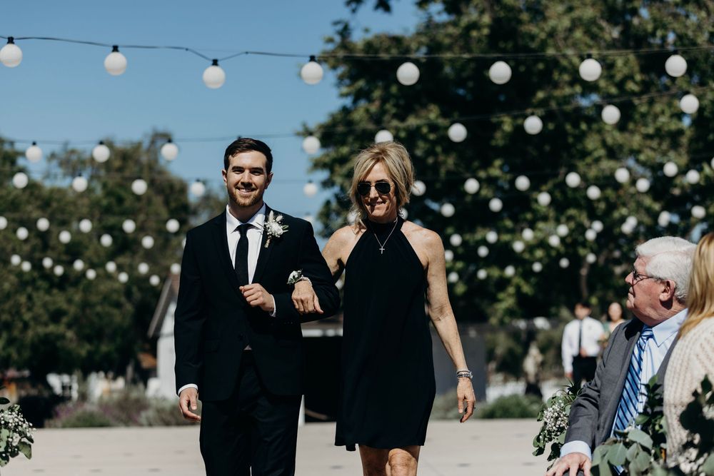 Wedding guests walk along an outdoor path decorated with string lights during a daytime celebration.