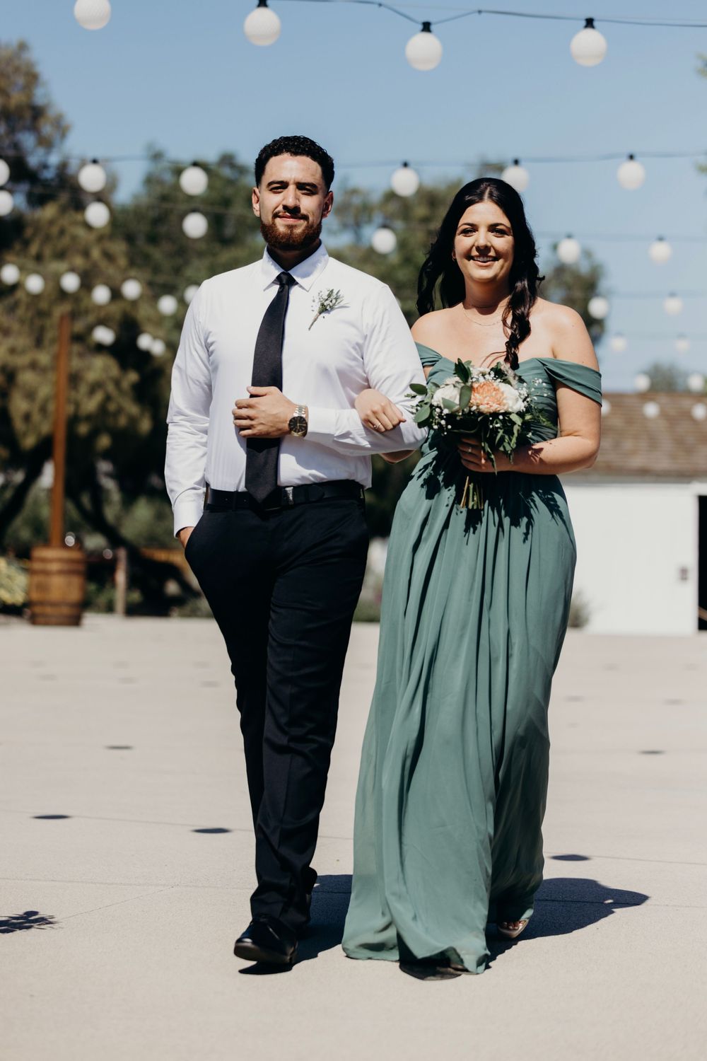 A couple in formal attire walking outdoors under string lights at a wedding celebration.