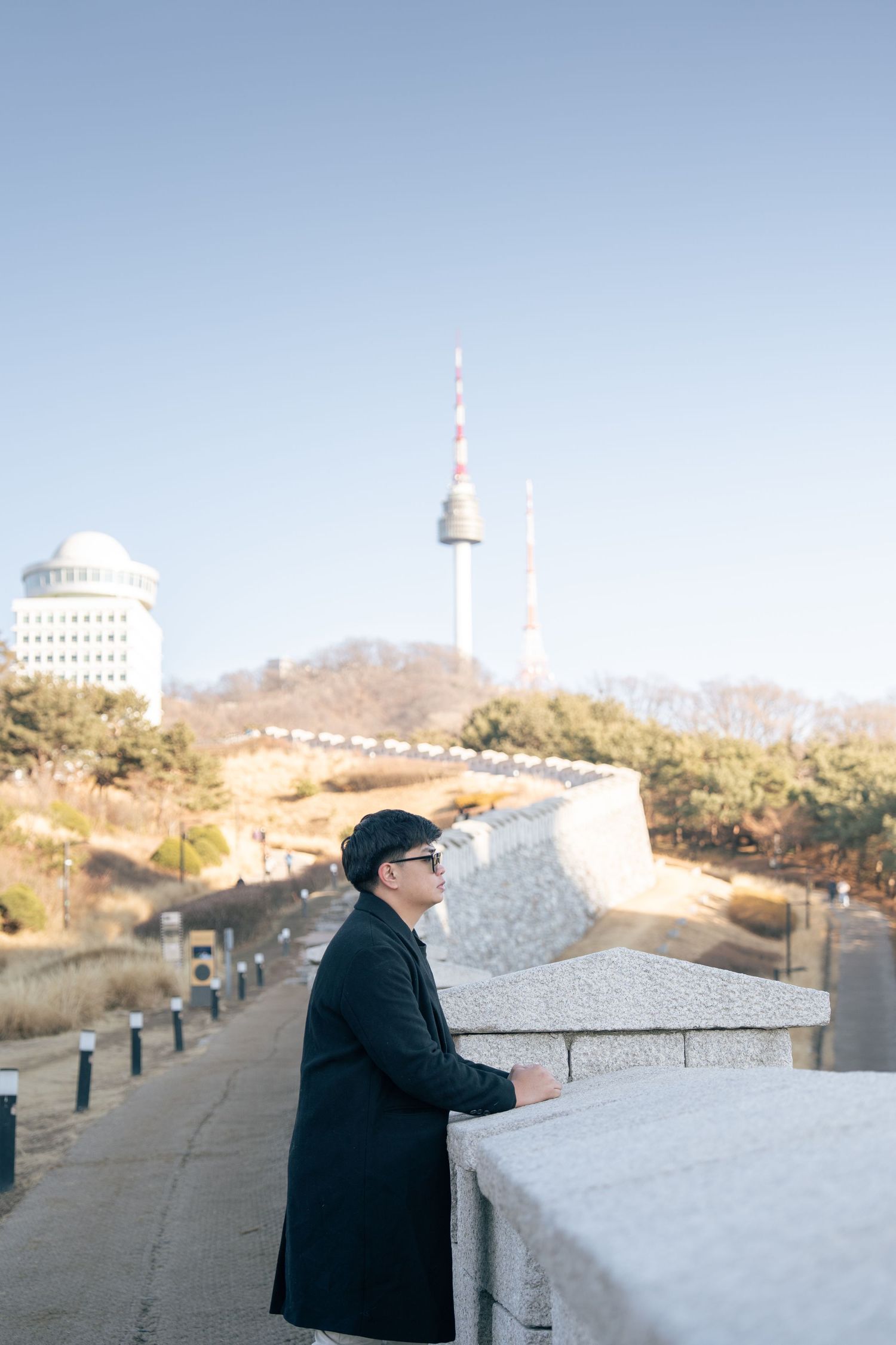 Person in black coat looking out over a scenic overlook with mountains and buildings in the distance.