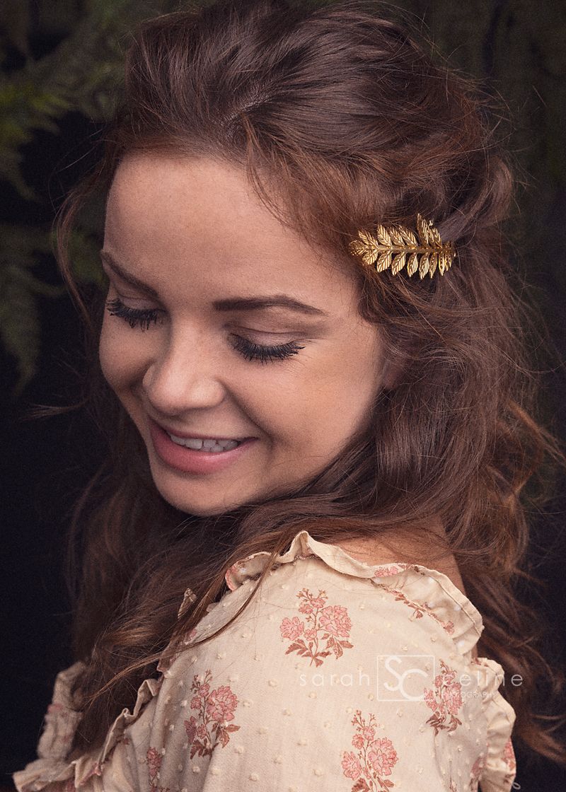 A person wearing a vintage-style cream dress with floral details and a delicate gold leaf hair accessory smiles warmly.