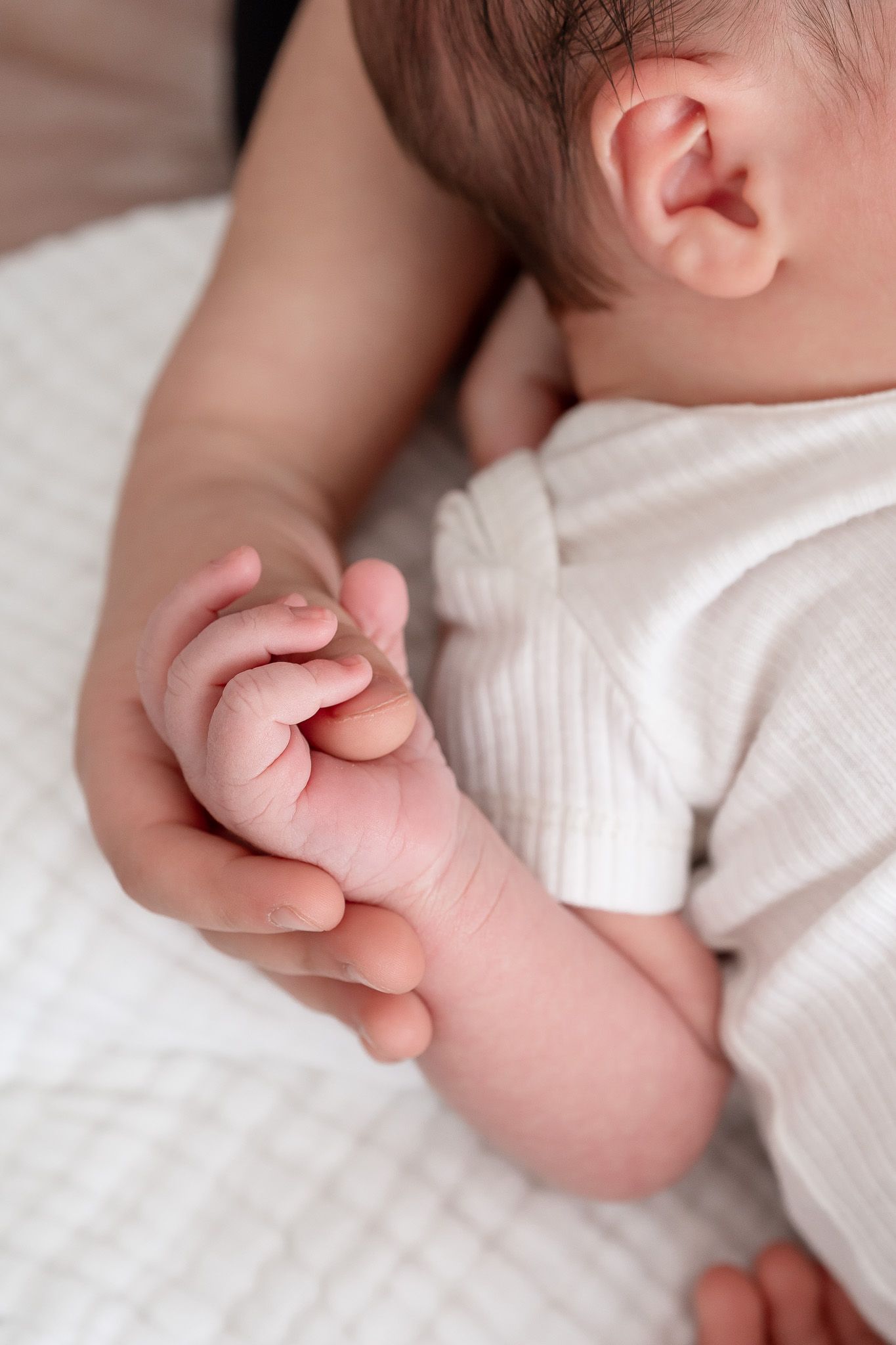 Close-up of a tender moment as a tiny baby's hand grasps an adult finger while wearing a white outfit.